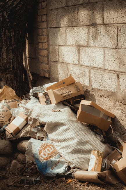 A large pile of empty wooden fruit crates, predominantly light-colored with visible wood grain textures, are stacked haphazardly in an outdoor area. The crates feature printed labels with images of fr